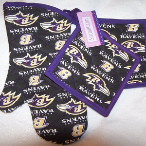 Op de afbeelding: Set van drie zwarte en paarse ovenwantjes met een gestikt ontwerp. De wantjes hebben een herhalend patroon van het Baltimore Ravens-logo, dat een ravenkop met een 'B' op de borst en de woorden "Baltimore Ravens" bevat. De wantjes zijn afgewerkt met paarse stof.