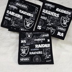 May include: Three square quilted pot holders featuring a black and white Las Vegas Raiders pattern. The fabric displays the Raiders team logo, the text Las Vegas, and graphic illustrations of the Las Vegas skyline, including a large Ferris wheel.