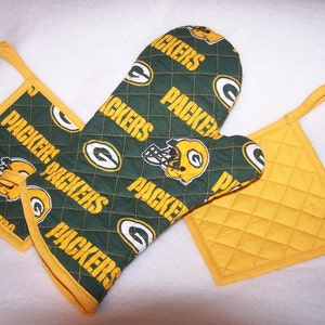 May include: Green and yellow oven mitt and pot holder set with the word "Packers" repeated on the fabric. The set is quilted and has a hanging loop.