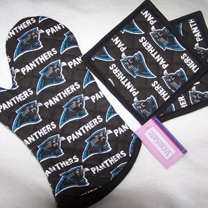 Op de afbeelding: Een set zwarte en blauwe ovenwanten en pannenlappen met het logo van de Carolina Panthers. De wanten en lappen zijn gestikt en hebben een zwarte rand. De tekst "PANTHERS" wordt herhaald op de stof.