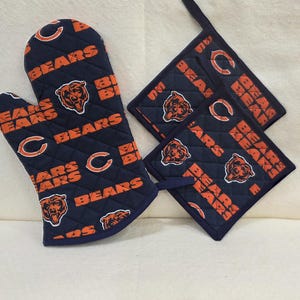 May include: A navy blue quilted oven mitt and two matching square pot holders featuring an orange and white Chicago Bears logo and the word BEARS printed in orange text throughout the fabric pattern, with dark blue trim on all edges.
