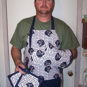 May include: A white and blue kitchen apron with a navy blue trim and a pattern of football helmets and the words "Texans" repeated. The apron is being worn by a person. There are also two matching pot holders with the same pattern.