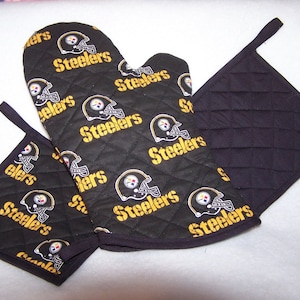 Pode incluir: Um conjunto de duas luvas de forno acolchoadas pretas e um descanso de panela com o logotipo do Pittsburgh Steelers e a palavra "Steelers" repetida em letras amarelas.