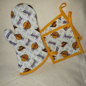 May include: Set of three Los Angeles Lakers themed kitchen pot holders. One is a large oven mitt and two are smaller pot holders. All are white with a yellow quilted pattern and feature the Lakers logo and basketballs.