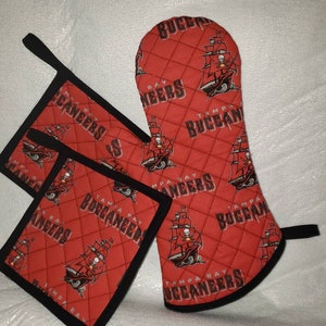 Op de afbeelding: Rode gewatteerde ovenwant, pannenlappen en vierkante onderzetters met zwarte rand. De items hebben een herhalend patroon van een piratenschip en de woorden "TAMPA BAY BUCCANEERS".