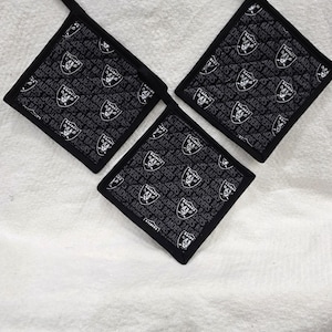 May include: Three square black fabric pot holders featuring a repeating white Raiders logo and the text Raiders printed throughout. Each pot holder has a black fabric loop attached to one corner for hanging, with black trim along the edges.