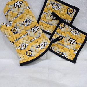 May include: A quilted oven mitt and two square pot holders featuring a yellow and white Nashville Predators hockey team pattern with black trim. The fabric displays the team logo, the text Nashville Predators, and the NP monogram throughout.