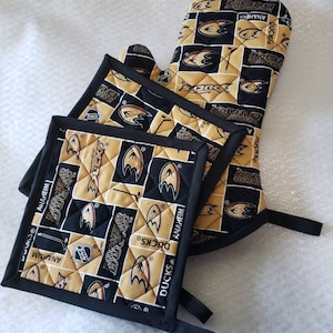 Op de afbeelding: Set van drie zwarte en gouden ovenwantjes met het logo van het NHL-team Anaheim Ducks. De wantjes zijn gewatteerd en hebben een ophanglus.