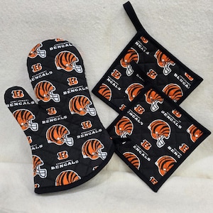 May include: A quilted oven mitt and two square pot holders in black fabric featuring an all-over print of orange and black striped football helmets and the text Cincinnati Bengals. Each piece has black trim and a hanging loop for storage.