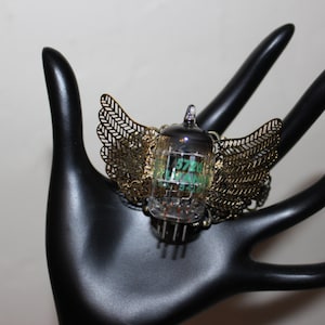 May include: A unique ring featuring a clear glass tube with green and white internal components, framed by ornate gold-colored metal wings. The ring is set on a black hand-shaped display, showcasing its intricate design.
