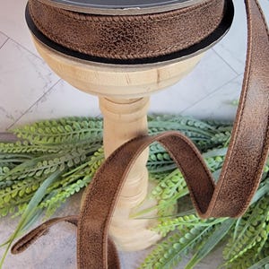 May include: A spool of brown ribbon with a textured, faux-leather appearance and stitched edges. The black spool features the text Lenore Arnold Designer Ribbon and Reliant Ribbon with decorative floral graphics printed in white on the top surface.
