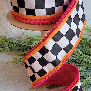 May include: A spool of wired ribbon featuring a black and white checkered pattern. The edges of the ribbon are trimmed with a bright orange fabric border and a decorative hot pink pom-pom fringe that runs along both sides of the fabric strip.