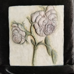 May include: A white ceramic tile with a raised relief design of three pink roses on a stem with green leaves.