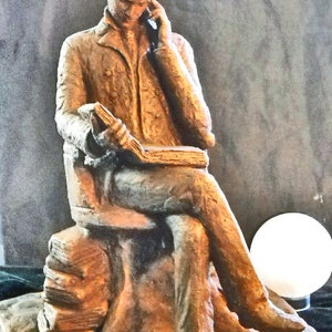 May include: A bronze sculpture of a person seated on a stone bench, holding a book and looking down. The sculpture is on a stone base.
