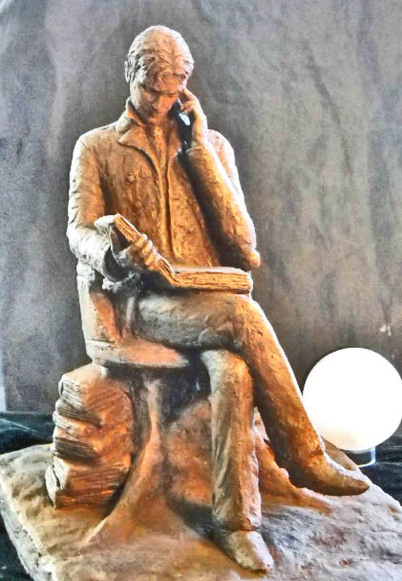 May include: A bronze sculpture of a person seated on a stone bench, holding a book and looking down. The sculpture is on a stone base.