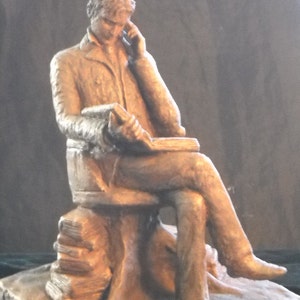 May include: A bronze sculpture of a person sitting on a stack of books, holding a book in their lap and looking down at it.