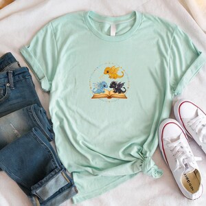 Pu&ograve; includere: T-shirt verde chiaro con un disegno circolare con tre draghi dei cartoni animati: oro, blu e nero, attorno a un libro aperto. La maglietta &egrave; abbinata a jeans blu e scarpe da ginnastica bianche.