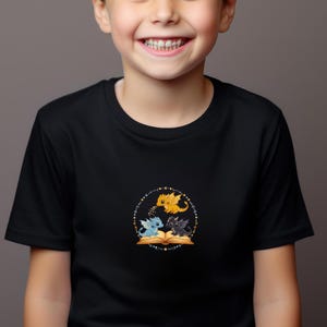 Pu&ograve; includere: T-shirt nera con un disegno circolare con tre draghi dei cartoni animati. Un drago giallo &egrave; sopra due draghi, uno blu e uno nero, riuniti attorno a un libro aperto. Il disegno &egrave; circondato da un cerchio tratteggiato con stelle.
