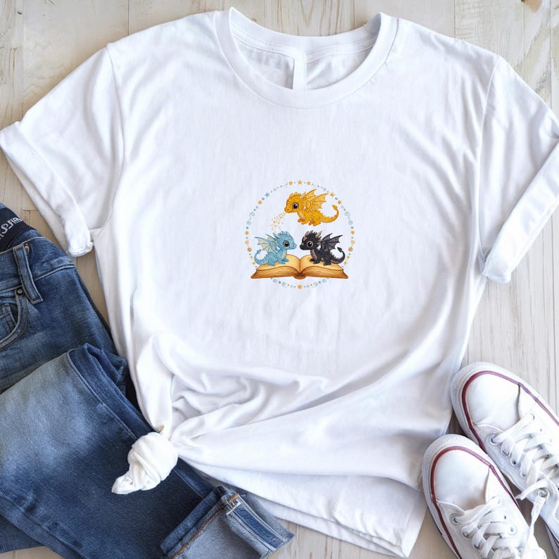 May include: White t-shirt featuring a whimsical design of three cartoon dragons. A yellow dragon hovers above two dragons, one blue and one black, who are sitting on an open book. The design is surrounded by a dotted circle.