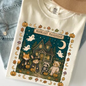 May include: A cream-colored t-shirt featuring a graphic of a haunted house and four costumed animals: a fox, a rabbit, a raccoon, and a bear. The text reads MOONLIGHT MASQUERADE. The border includes pumpkins, leaves, moons, and mushrooms.