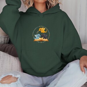 Pu&ograve; includere: Felpa con cappuccio verde scuro con un disegno circolare di tre draghi dei cartoni animati: giallo, blu e nero. Sono posizionati attorno a un libro aperto, all'interno di un cerchio punteggiato con stelle. La felpa ha una tasca a marsupio e un cappuccio.