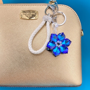 Puede incluir: Un bolso dorado con un llavero decorativo. El llavero presenta una correa trenzada blanca, un acento plateado brillante y una flor de cristal azul. El bolso tiene una placa metálica dorada con el nombre de la marca.