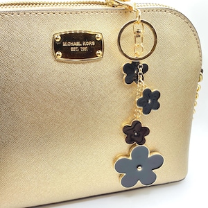 May include: A gold-colored Michael Kors handbag with a gold zipper and a decorative flower charm. The charm features black flower-shaped pendants hanging from a gold ring. The Michael Kors logo is visible on the front of the bag.