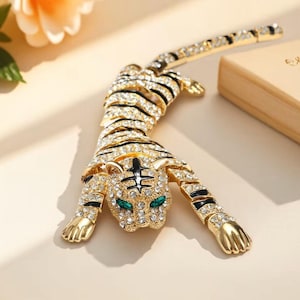 Può includere: Ciondolo a forma di tigre dorata incrostato di cristalli trasparenti e strisce di smalto nero. Gli occhi della tigre sono verde smeraldo. Il ciondolo è in posa strisciante, con le zampe distese.