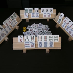 May include: A Mahjong set with wooden tile racks and dice. The tiles are white with black, red, and blue symbols and characters. The set is arranged on a dark surface, ready for play.