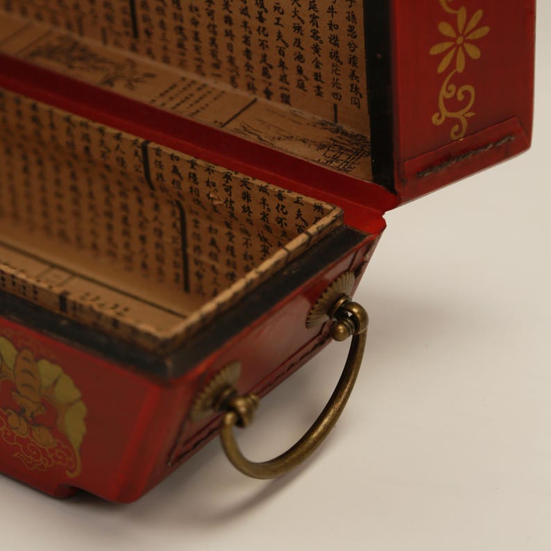 May include: A red wooden box with a hinged lid, gold floral designs and a brass handle. The interior is lined with paper printed with Asian characters. The box is open, revealing the inside.