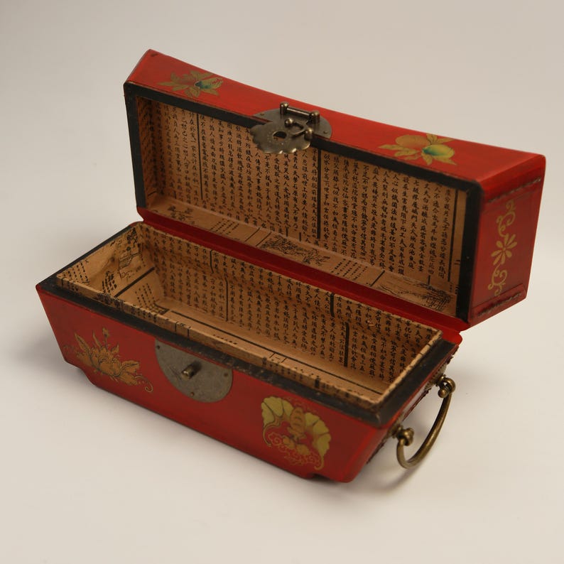 May include: A red rectangular box with a hinged lid, open to reveal an empty interior lined with printed paper. The box is decorated with gold floral designs and a metal clasp. A metal handle is attached to the side.