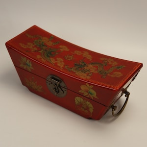 May include: A red wooden box with a curved top, decorated with a dragon and floral design in gold and green. The box has a metal clasp and handle, reflecting an Asian aesthetic. The box is approximately 30 cm long.