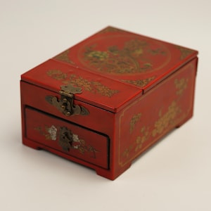 Puede incluir: Una caja de joyería de madera roja con tapa con bisagras y un pequeño cajón. La caja está decorada con diseños florales dorados y un patrón circular en la tapa. Cuenta con un cierre y bisagras de latón.