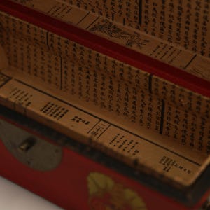 May include: Open red wooden box with a gold clasp. The interior is lined with aged paper featuring black Chinese characters. The box's edges are red, and the interior paper has a faded, antique appearance.
