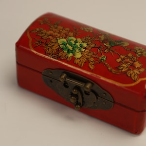 Könnte beinhalten: Kleine, rote Holzkiste mit gewölbtem Deckel und Metallverschluss. Die Kiste ist mit einem Blumen- und Vogeldesign in Gold, Grün und Gelb verziert. Die Kiste ist geschlossen.