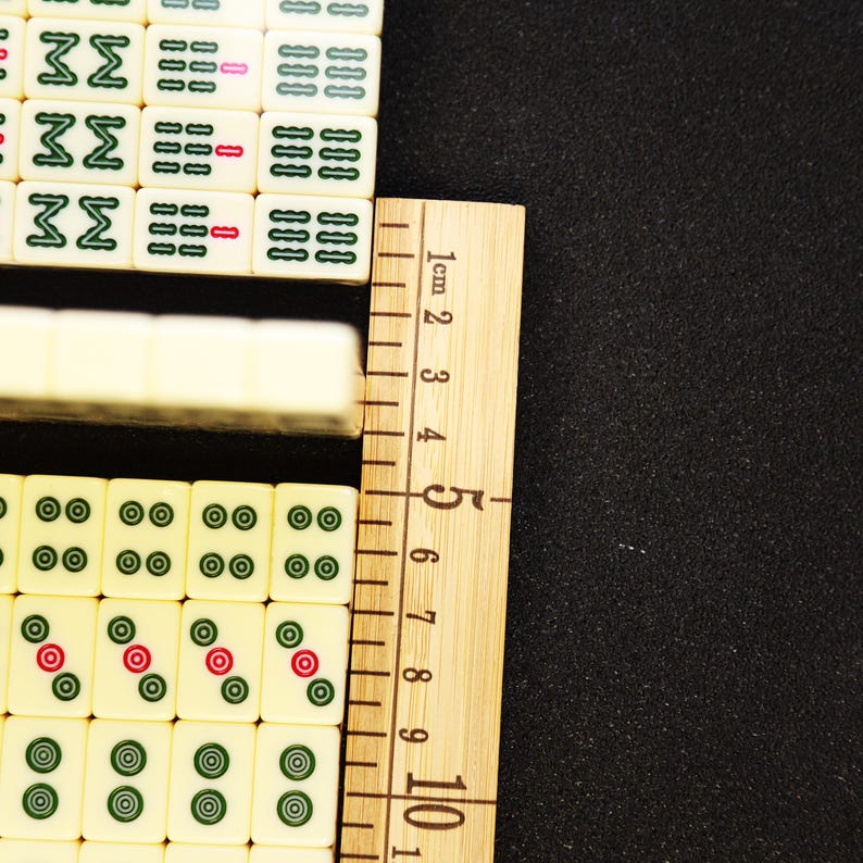 May include: Close-up of ivory-colored Mahjong tiles with green and red circular markings. A wooden ruler with centimeter markings is positioned next to the tiles. The ruler measures up to 10 cm.