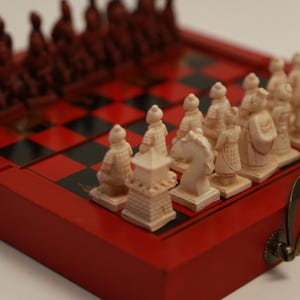 May include: A red and black chessboard with chess pieces. The chess pieces are carved in the shape of ancient warriors and are in shades of red and cream. The board is red with black squares.