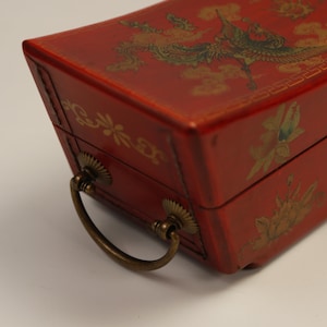May include: A red rectangular box with a brass handle and gold detailing. The box is decorated with a painted bird and floral design. The handle is curved and attached with decorative hardware.