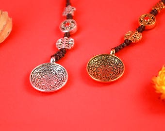 Puede incluir: Dos colgantes decorativos con diseños intrincados cuelgan sobre un fondo rojo. Uno es plateado, el otro dorado, ambos con patrones detallados. Los colgantes están sujetos a cordones marrones oscuros con cuentas decorativas.