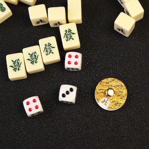 May include: Overhead shot of Mahjong game pieces on a dark surface. The ivory-colored tiles have green Chinese characters. Dice with red and black dots are scattered around a gold-colored coin-shaped piece.