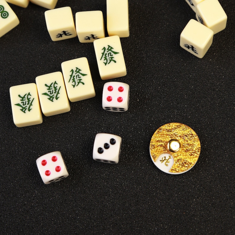 May include: Overhead shot of Mahjong game pieces on a dark surface. The ivory-colored tiles have green Chinese characters. Dice with red and black dots are scattered around a gold-colored coin-shaped piece.