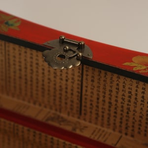 May include: Close-up of a red wooden box with a decorative brass clasp. The box features a floral design and is lined with pages of text. The clasp is ornate, adding a vintage aesthetic.