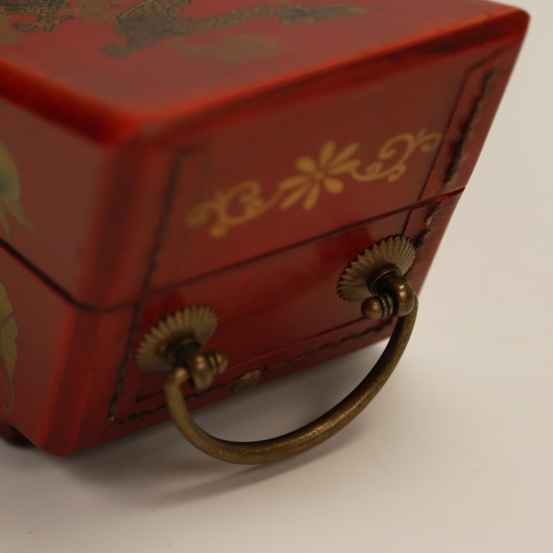 May include: A red lacquered box with gold floral and dragon designs. The box has a trapezoidal shape and features two brass handles. The box is likely used for jewelry or small keepsakes.