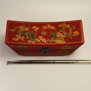 May include: A red wooden box with a curved lid, featuring gold and green dragon and phoenix designs. The box has a metal clasp and decorative border. The box measures approximately 30 cm long.