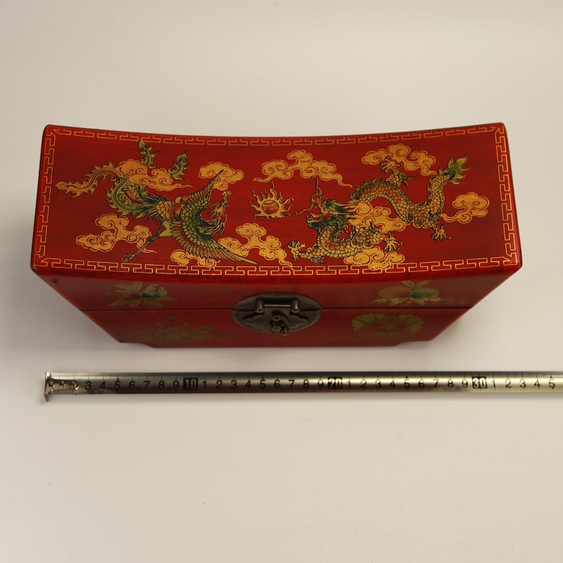 May include: A red wooden box with a curved lid, featuring gold and green dragon and phoenix designs. The box has a metal clasp and decorative border. The box measures approximately 30 cm long.