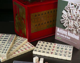 May include: A complete Mahjong set featuring ivory-colored tiles with red and green markings, dice, and a red box with gold accents. A handbook titled "Mahjong Handbook" is also included. The set is designed for playing the classic Chinese tile game.