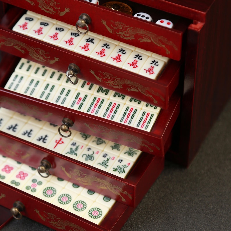 May include: A red wooden Mahjong set with multiple drawers, each filled with ivory-colored tiles. The drawers are adorned with gold dragon designs and brass hardware. The tiles feature Chinese characters and colorful symbols.