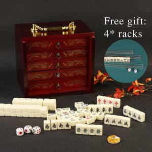 May include: A complete Mahjong set with a dark red wooden storage box featuring five drawers with gold dragon designs. The set includes ivory-colored tiles, dice, and a small dish. The image also includes the text "Free gift: 4* racks."