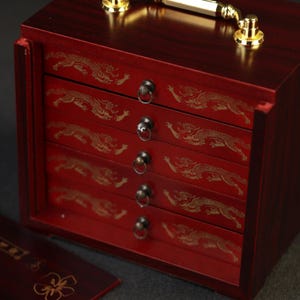 May include: A red wooden jewelry box with four drawers, each adorned with a gold dragon design. The box has a gold handle and hardware. The drawers have small, round metal pulls.