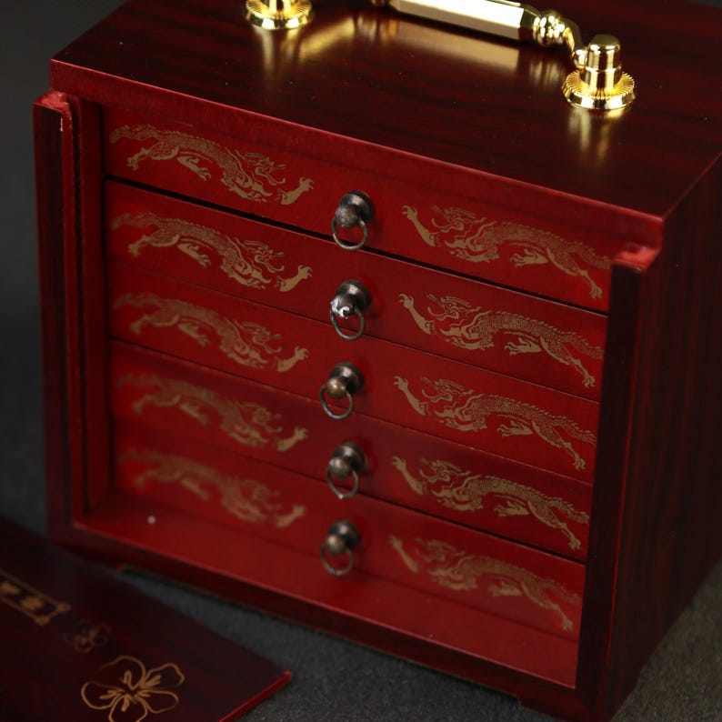 May include: A red wooden jewelry box with four drawers, each adorned with a gold dragon design. The box has a gold handle and hardware. The drawers have small, round metal pulls.